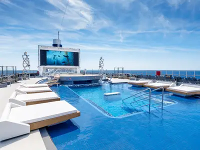 Blick auf den weitläufigen Long Island Pool der MSC Seashore. Mehrere weisse Sonnenliegen stehen im und am Pool, im Hintergrund eine grosse Leinwand mit Unterwasseraufnahmen. Blauer Himmel über dem Meer.