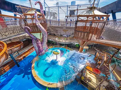 Blick von oben auf den bunten Pirates Cove Aquapark an Bord der MSC Seashore. Ein riesiger lila Oktopus ragt über eine Wasserrutsche, während Wasser aus einem großen Eimer in ein Becken stürzt. Weitere Rutschen und ein Kletternetz sind sichtbar.
