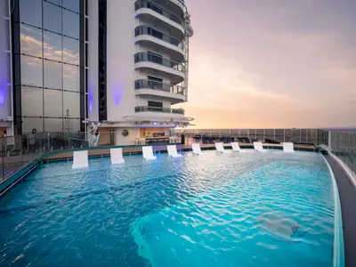 Der Infinity Pool an Bord der MSC Seashore bei Sonnenuntergang. Weiße Liegen im blauen Wasser, eine Bar und die spiegelnde Glasfassade des Schiffs. Rechts ist das offene Meer zu sehen.
