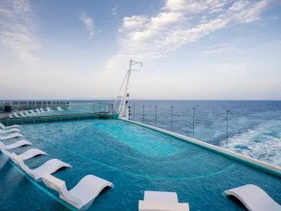 Ein weitläufiger Infinity Pool an Bord der MSC Seashore mit weißen Liegen im und am Wasser. Im Hintergrund das weite blaue Meer und Gischt.