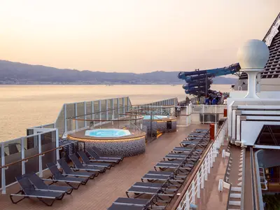 Sonnendeck der MSC Seashore im Abendlicht. Whirlpools, eine Wasserrutsche und Liegestühle sind zu sehen. Blick auf das Meer und ferne Berge.