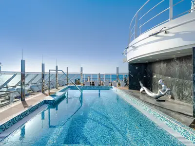 Ein sonniges Pooldeck an Bord der MSC Seashore. Ein blauer Mosaikpool und eine silberne Meerjungfrauen-Skulptur sind im Vordergrund zu sehen. Im Hintergrund ist das Meer sichtbar, umgeben von eleganten Glasgeländern und Liegestühlen.