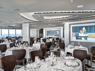 Ein elegantes, helles Restaurant an Bord der MSC Seashore mit vielen gedeckten Tischen, bequemen braunen Stühlen und großen Panoramafenstern mit Blick auf das Meer. Die Decke ist geschwungen mit integrierten Lichtinstallationen, und an den Wänden hängen moderne Kunstwerke. Es sind keine Personen zu sehen.