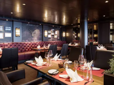 Elegant beleuchtetes Spezialitätenrestaurant Butcher's Cut an Bord der MSC Seashore. Gedeckte Tische, rote Polsterbänke, dunkle Wände mit Rindermotiven. Ohne Personen.