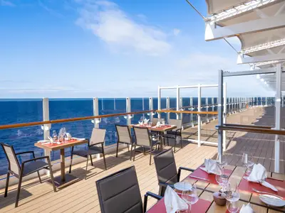 Outdoor-Restaurant an Bord der MSC Seashore mit gedeckten Tischen. Blick auf das weite, blaue Meer unter klarem Himmel. Moderne Einrichtung mit Glasgeländern.