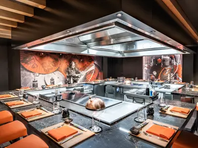 Das Kaito Teppanyaki Restaurant an Bord der MSC Seashore. Moderne Kochstationen mit Marmortischen, orangefarbenen Servietten und Ledersesseln. Eine grosse Abzugshaube und japanische Wandbilder.