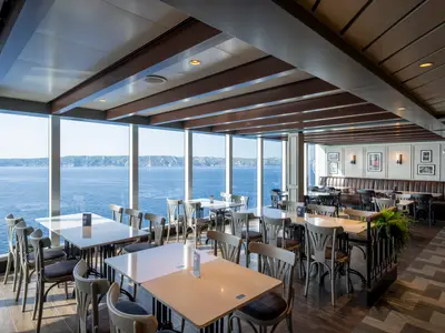 Helles Restaurant an Bord der MSC Seashore mit grossen Fenstern und weitem Meerblick auf die Küste. Moderne Einrichtung mit Tischen, Stühlen und Sitzbänken. Ohne Personen.