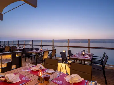 Romantisches Aussenrestaurant der MSC Seashore bei Sonnenuntergang. Gedeckte Tische mit roten Sets und Kerzen. Blick auf das ruhige Meer und den Dämmerungshimmel. Eine einladende Atmosphäre.