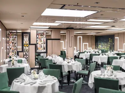 Das elegant gedeckte Manhattan Restaurant an Bord der MSC Seashore. Zahlreiche Tische mit weissen Tischtüchern und grünen Stühlen füllen den Raum, beleuchtet von Deckenlichtern und hinterleuchteten Wänden. Im Hintergrund eine grosse Stadtsilhouette an der Wand und dekorative Weinregale.