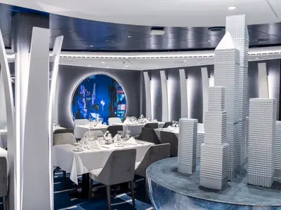 Das moderne 5th Avenue Restaurant an Bord der MSC Seashore. Elegante Tische mit weißer Wäsche, grauen Stühlen, einem runden Skyline-Mural und weißen Wolkenkratzer-Skulpturen schmücken den Raum.