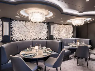 Ein modernes Spezialitätenrestaurant an Bord der MSC Seashore. Geschwungene graue Lederbänke umrunden runde Tische, die für ein gehobenes Abendessen eingedeckt sind. Die Wände sind mit auffälligen, wellenartigen weißen Dekorationen verziert. Zwei große, gläserne Kronleuchter spenden warmes Licht.
