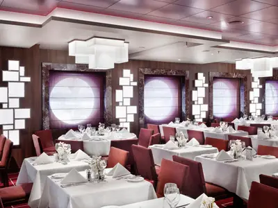 Ein elegantes Restaurant an Bord der MSC Seashore mit vielen gedeckten Tischen, roten Stühlen, modernen Leuchten und großen Fenstern mit violetten Rollos. Die Einrichtung ist modern und einladend.