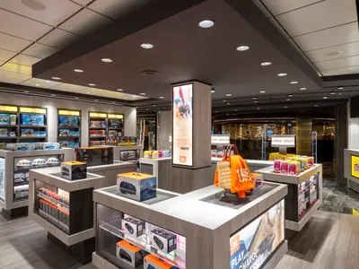 Innenansicht eines modernen Duty-Free-Shops an Bord der MSC Seashore. Regale sind mit Lego-Sets gefüllt, Auslagen präsentieren JBL-Lautsprecher und Süßigkeiten wie M&M's. Ein großer beleuchteter Pfeiler zeigt Werbung für JBL-Produkte.