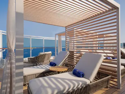 Luxuriöses Sonnendeck an Bord der MSC Seashore. Elegante Lounge-Stühle stehen unter Holzpergolen mit weitem Blick auf das blaue Meer. Blaue Handtücher liegen auf den weißen Polstern bereit.