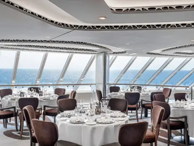 Das exklusive MSC Yacht Club Restaurant an Bord der MSC Seashore. Elegante Tische mit weißen Decken und braunen Stühlen stehen vor großen Fenstern mit Panoramablick auf das Meer.