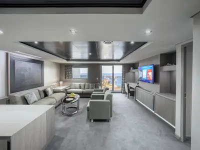 Das luxuriöse Wohnzimmer einer Suite an Bord der MSC Seashore. Graue Möbel, ein Glastisch und ein großer Fernseher. Große Fenster und eine Balkontür bieten Meerblick.
