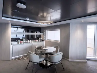 Modernes Wohnzimmer einer luxuriösen Owner's Suite an Bord der MSC Seashore. Es zeigt einen runden Marmortisch mit vier Stühlen, eine Barzeile mit Kaffeeautomat und Getränken sowie große Fenster zu einem Balkon.