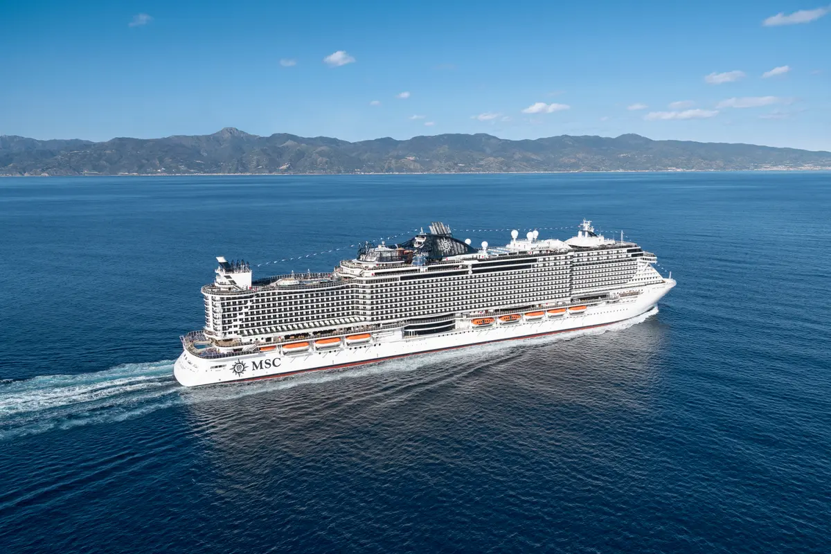 Luftaufnahme der MSC Seaside, die auf dem offenen Meer fährt und eine weiße Heckwelle hinter sich zieht. Im Hintergrund sind grüne Berge unter blauem Himmel sichtbar.