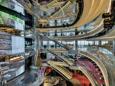 Das luxuriöse, mehrstöckige Atrium der MSC Seaside mit einer großen LED-Wand, die einen Wasserfall zeigt. Gläserne, beleuchtete Treppen und elegante Bars sind sichtbar, im Hintergrund das Meer.