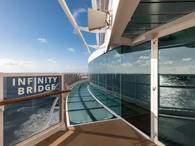 Die Infinity Bridge an Bord der MSC Seaside. Die gläserne Brücke und Balustrade bieten einen weiten Blick über das Meer unter blauem Himmel. Sonnenlicht spiegelt sich im Wasser. Keine Personen.