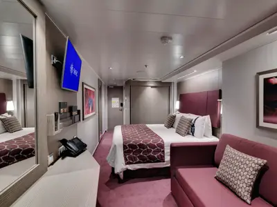 Eine modern eingerichtete Balkonkabine an Bord der MSC Seaside. Die Kabine ist mit einem Doppelbett, einem Sofa und einem Fernseher ausgestattet. Dominante Farben sind Weinrot und Grau. Ein Telefon steht auf einem Schreibtisch.