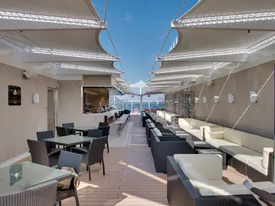 Aurea Bar an Bord der MSC Seaside: Helle, ueberdachte Aussenbar mit Theke, Hockern und Rattan-Sitzlounges. Sonnensegel spenden Schatten. Im Hintergrund das weite blaue Meer.