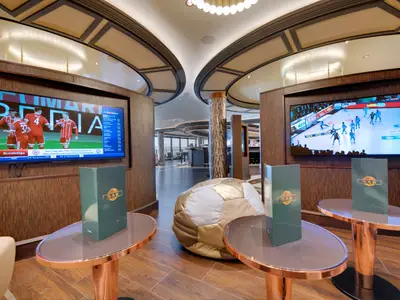 Innenansicht der Sports Bar an Bord der MSC Seaside mit zwei grossen Bildschirmen, die Sportevents zeigen. Moderne Holzvertäfelung, runde Tische und ein Fussball-Sitzsack sind zu sehen.