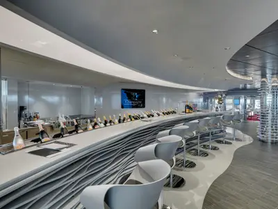 Ansicht der eleganten Champagne Bar an Bord der MSC Seaside mit langer, geschwungener Theke, vielen Champagnerflaschen und modernen Barhockern. Ein beleuchteter Bildschirm zeigt den Schriftzug 'Champagne Bar'.