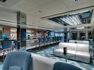 Blick in die schicke Wine & Cocktails Bar an Bord der MSC Seaside mit langer Theke, blauen Barhockern und reflektierenden Decken. Sitzgelegenheiten mit weissen Ledersofas und kleinen Tischen sind im Vordergrund zu sehen.