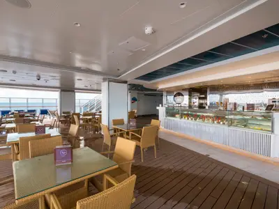 Die Venchi 1878 Gelato & Crêperie an Bord der MSC Seaside. Der helle Innenraum bietet zahlreiche Tische und Stühle, große Fenster mit Meerblick und eine lange Verkaufstheke mit Dessertauslage. Der Boden ist teilweise aus Holz, teilweise aus Marmor.