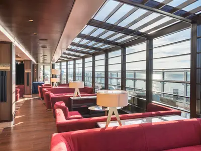 Die Seaview Lounge an Bord der MSC Seaside zeigt eine moderne, tageslichtdurchflutete Bar-Lounge mit roten Sitzgelegenheiten, Tischen und Lampen, umgeben von großen Panoramafenstern mit Blick auf das Meer.