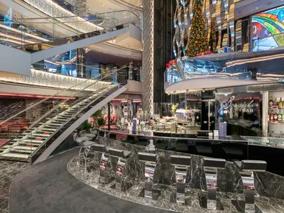 Elegantes Atrium der MSC Seaside. Im Vordergrund die Seaside Bar mit Marmortheke und Hockern. Eine breite Treppe, ein Weihnachtsbaum und bunte Glasfenster sind sichtbar.