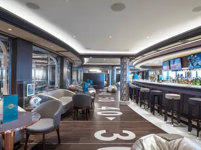 Blick in die sportlich thematisierte Sports Bar an Bord der MSC Seaside. Im Vordergrund ein langer Bartresen mit Stühlen, dahinter Lounge-Sitzbereiche. Der Boden ist wie ein Sportfeld markiert. Sport-Memorabilien in Vitrinen.