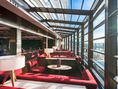 Innenansicht der Seaview Lounge an Bord der MSC Seaside. Der helle Raum bietet bequeme rote Sofas und Tische. Grosse Fenster und Glasdach mit Meer- und Hafenblick.