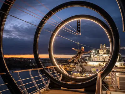 Ein Mann gleitet lachend an einer Zip-Line durch beleuchtete Ringe an Deck der MSC Seaside bei Sonnenuntergang. Im Hintergrund die Skyline einer Stadt und der Hafen.