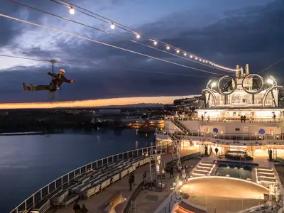 Ein Mann schwebt an einer Zip-Line über dem oberen, beleuchteten Deck der MSC Seaside bei Sonnenuntergang. Im Hintergrund sind eine Küstenstadt und Berge erkennbar, das Meer spiegelt das Himmelslicht wider.