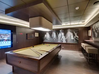 MSC Seaside: Eleganter Billiards-Salon mit Billardtisch, dunkler Holzvertäfelung, Art-déco-Wandkunst und einem Info-Bildschirm. Warme Beleuchtung.