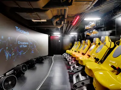 Ein interaktives XD-Kino an Bord der MSC Seaside. Mehrere Reihen leuchtend gelber Sitze sind auf einen großen Bildschirm ausgerichtet, der „Triotech – Creators of Fun“ zeigt. Ventilatoren stehen am Boden.