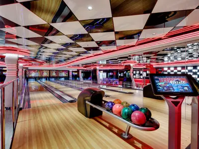Eine moderne Bowlingbahn mit drei Bahnen an Bord der MSC Seaside. Bunte Bowlingkugeln liegen bereit, umrahmt von roten Neonlichtern und einem Schachbrettmuster an der Decke.