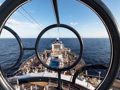 Blick vom Heck der MSC Seaside auf das offene Meer, eingerahmt von der Zip-Line-Struktur. Auf dem Sonnendeck entspannen zahlreiche Gaeste am Pool und auf Liegen unter blauem Himmel.