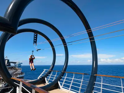 Eine Person mit Helm und Sicherheitsgeschirr fährt an der Zip Line über das Oberdeck der MSC Seaside. Im Hintergrund sind das Meer und der blaue Himmel zu sehen.