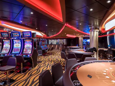 Modernes Casino der MSC Seaside mit Spielautomaten und Tischen für Blackjack/Roulette. Rote LED-Lichter an Decke, gemusterter Teppich und Barhocker prägen das Ambiente.
