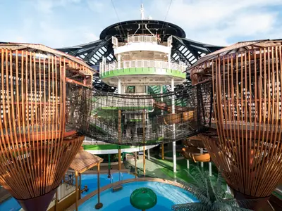Der Forest Aquaventure Park auf der MSC Seaside mit großem Netz-Kletterparcours, orangefarbenen Wasserrutschen und Pools. Die Brücke des Schiffes ist im Hintergrund unter blauem Himmel sichtbar.