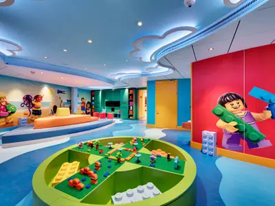 Der lebhafte LEGO-Spielbereich im Mini Club der MSC Seaside. Grosse LEGO-Figuren an den Waenden, ein runder Bautisch in der Mitte. Blauer Wellenboden, wolkenfoermige Lichter.