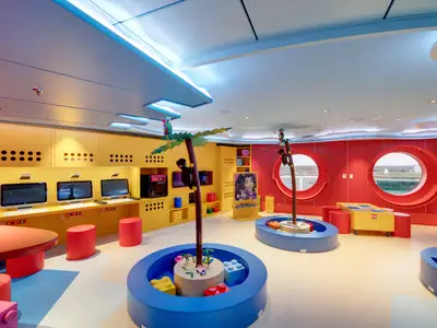 Der bunte Junior Club an Bord der MSC Seaside zeigt Spielbereiche mit Lego-Elementen, Computer-Stationen und rote Bullaugenfenster. Einladender Bereich für Kinder.