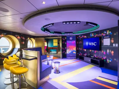 Der Young Club an Bord der MSC Seaside zeigt eine moderne Lounge mit runden Fenstern, Blick auf das Meer, einer Bar mit gelben Hockern und einer grossen MSC-Anzeige. Im Hintergrund ist ein Foosball-Tisch zu sehen.