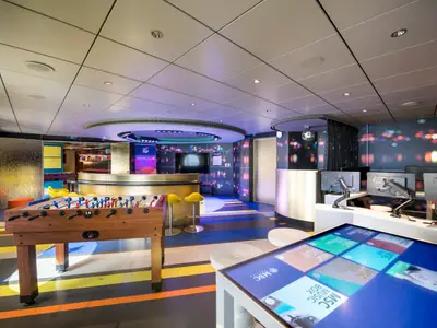 Der farbenfrohe Young Club an Bord der MSC Seaside zeigt einen Tischkicker, interaktive Touchscreens und Sitzgelegenheiten in einer lebhaften Umgebung mit bunten Lichtern und Mustern.