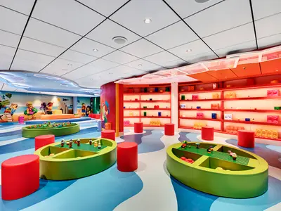 Der lebhafte LEGO Mini Club an Bord der MSC Seaside. Grüne Spieltische mit LEGO-Bausteinen und rote Hocker sind sichtbar. Die Wände zeigen bunte LEGO-Figuren und Regale mit großen LEGO-Steinen.