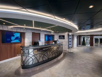 Die MSC Excursions Rezeption auf der MSC Seaside mit hellem Marmortresen, digitalen Anzeigetafeln, Holzpaneelen und einem festlichen Weihnachtsbaum in der Halle.