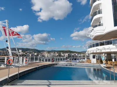 Der sonnige Aussenpool der MSC Seaside mit blauem Wasser. Im Hintergrund eine Hafenstadt mit Bergen und blauem Himmel. Rechts Schiffsdecks, links Flaggenmast.